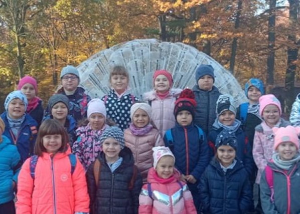 Grupa przedszkolaków w zimowych kurtkach i czapkach uśmiecha się do zdjęcia podczas jesiennej wycieczki w parku. W tle widać jesienne drzewa.