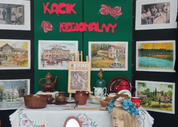 Wystawa „Kącik regionalny” ze starymi zdjęciami i obrazami na tablicach. Na stole z haftowanym obrusem stoją ceramiczne naczynia, dzbanki, książka „Biblia Ślązoka” i głowa z wiankiem