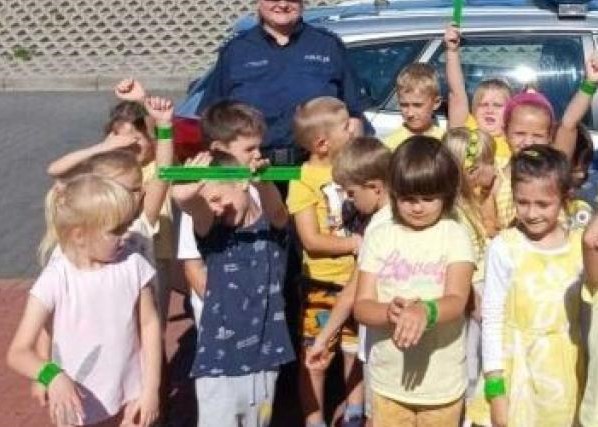 Grupa radosnych przedszkolaków z zielonymi odblaskowymi opaskami na rękach pozuje do zdjęcia z policjantem na tle radiowozu. Dzieci unoszą ręce w geście radości